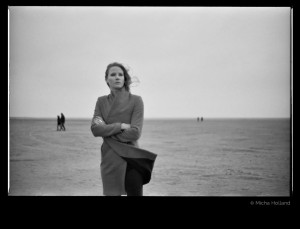 Woman with coat on the beach by Micha Holland. The photo shows Cate Martin wearing a coat. With crossed arms she is standing on the beach staring into the distance.